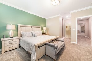 Quarto com cama, mesas de cabeceira, luminária, um banquinho e porta que dá para outro cômodo.