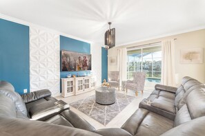 A living room with a blue accent wall, a large TV, leather sofas, a patterned armchair, and a view of a swimming pool. - Orlando Family Paradise with Private Pool & Resort (Four Corners)