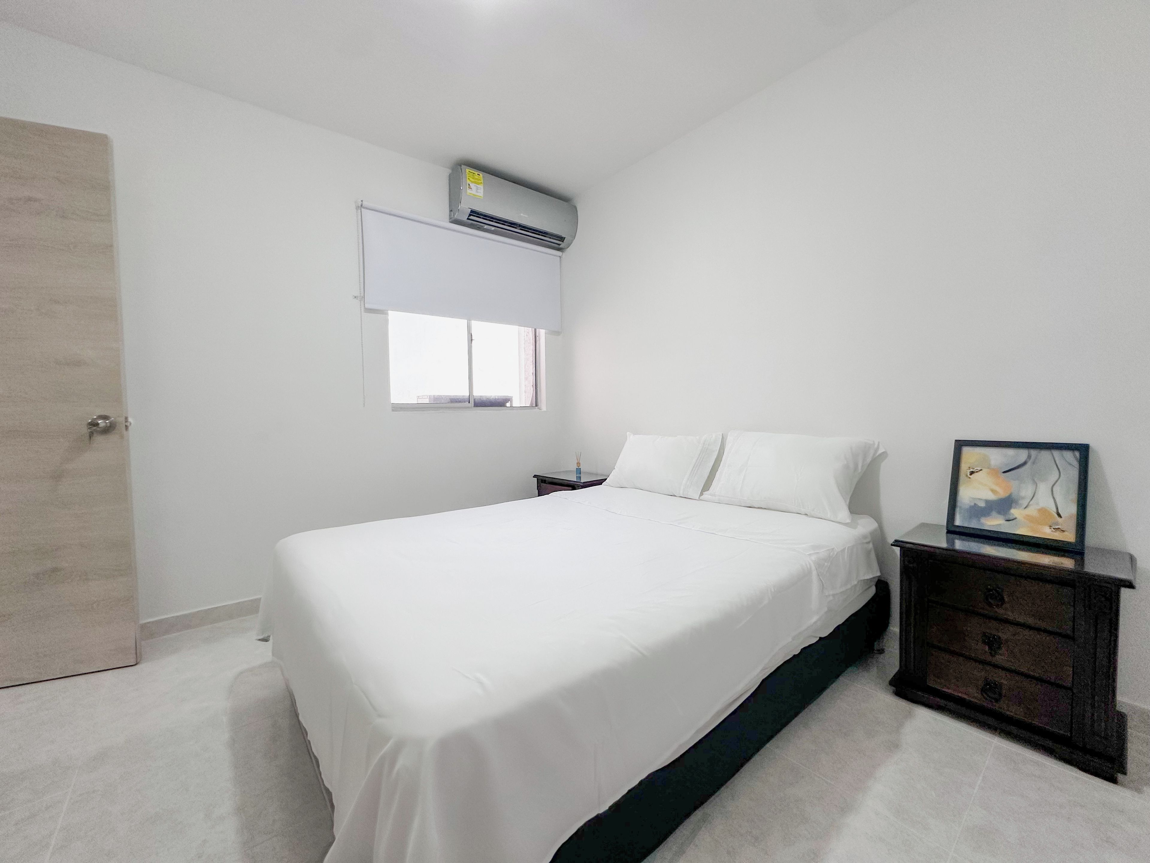 Un dormitorio con cama, mesita de noche, cómoda, unidad de aire acondicionado y ventana.