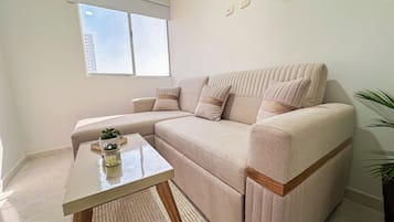 Un salon moderne avec un canapé d’angle beige, une petite table basse agrémentée d’une plante et une fenêtre avec des stores.