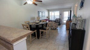 A dining area with a round table, chairs, and a ceiling fan. There is a living area with a couch and a television. The room has tiled flooring and a window with blinds. - 2BR Half Block to Beach, Garage, Heated Pool (South Padre Island)