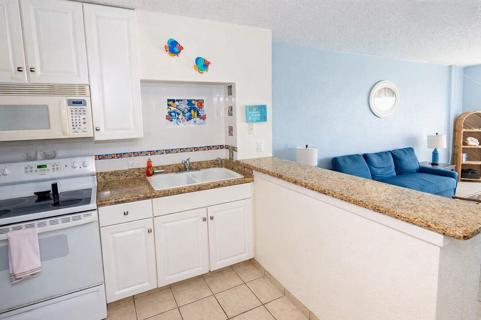 A compact kitchen with white cabinets, a microwave, stove, and a sink. There is a blue wall with a round mirror and a blue sofa in the adjacent living area.