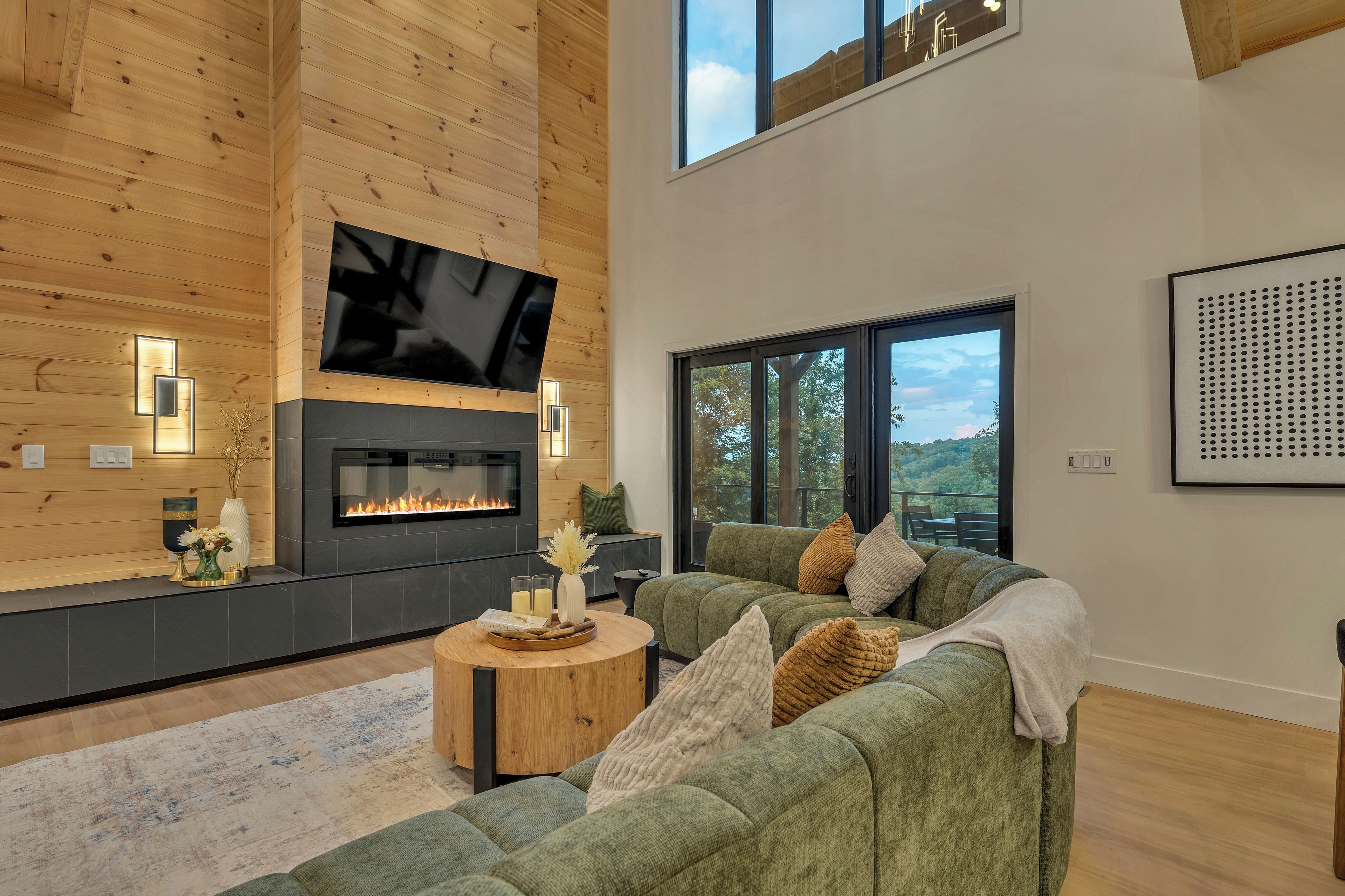 A modern living room with a fireplace, a flat-screen TV, and a large window offering a view of the outdoors.