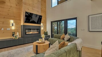 A modern living room with a fireplace, a flat-screen TV, and a large window offering a view of the outdoors.