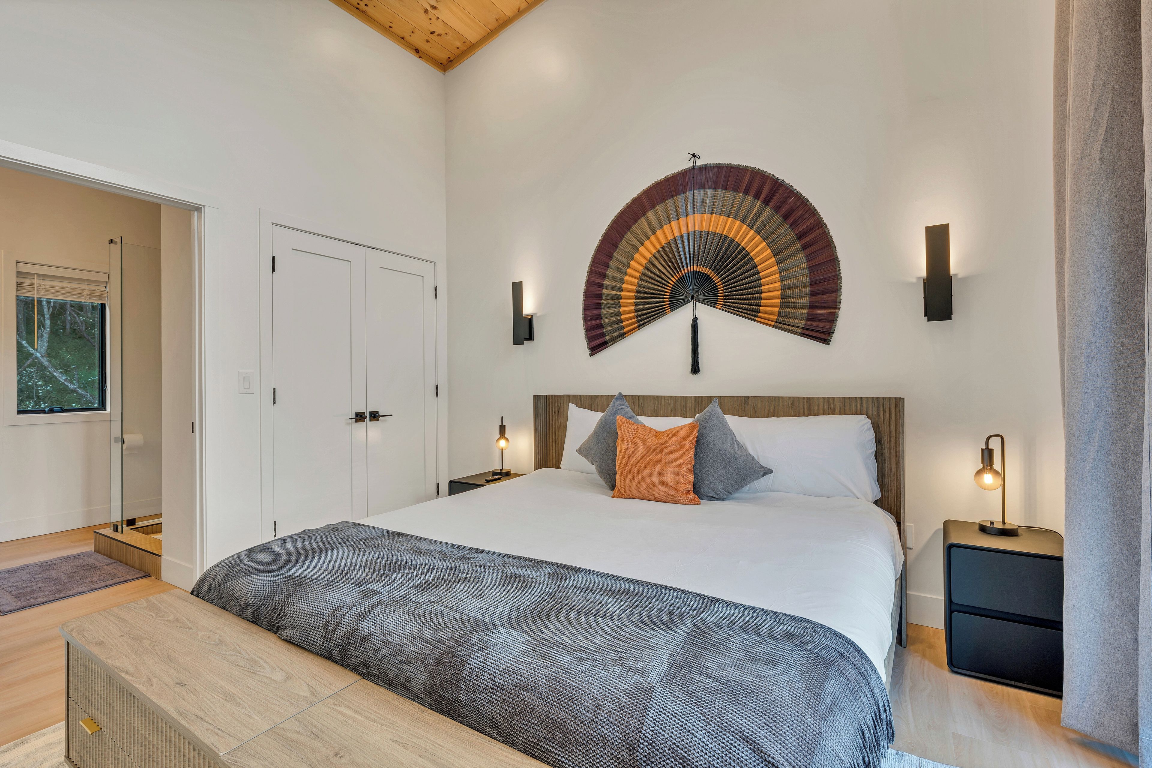 A modern bedroom with a large bed, bedside lamps, a wall-mounted fan, and a door leading to another room.