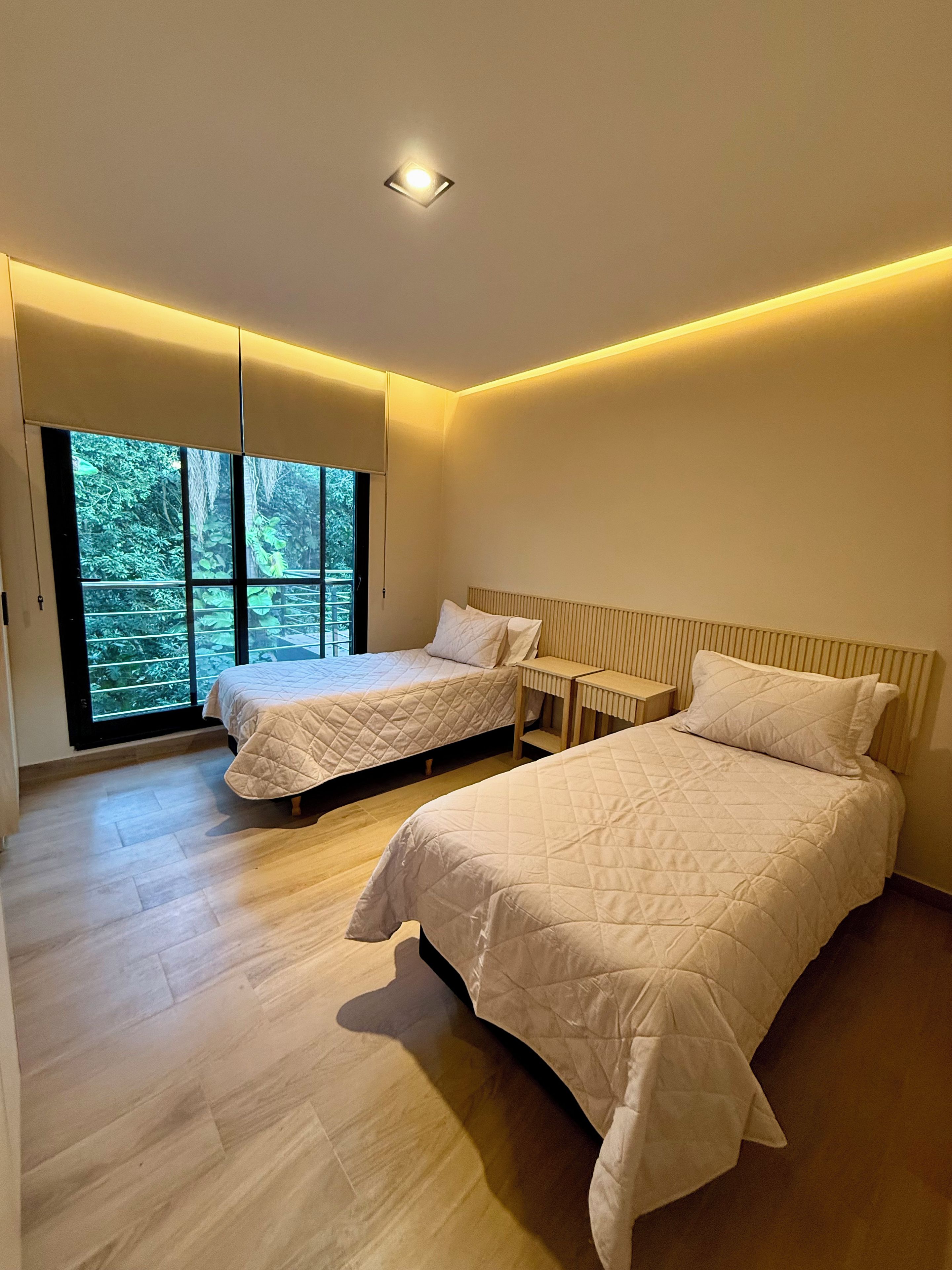 A modern bedroom with two beds, a window with a view of greenery, and a ceiling light.
