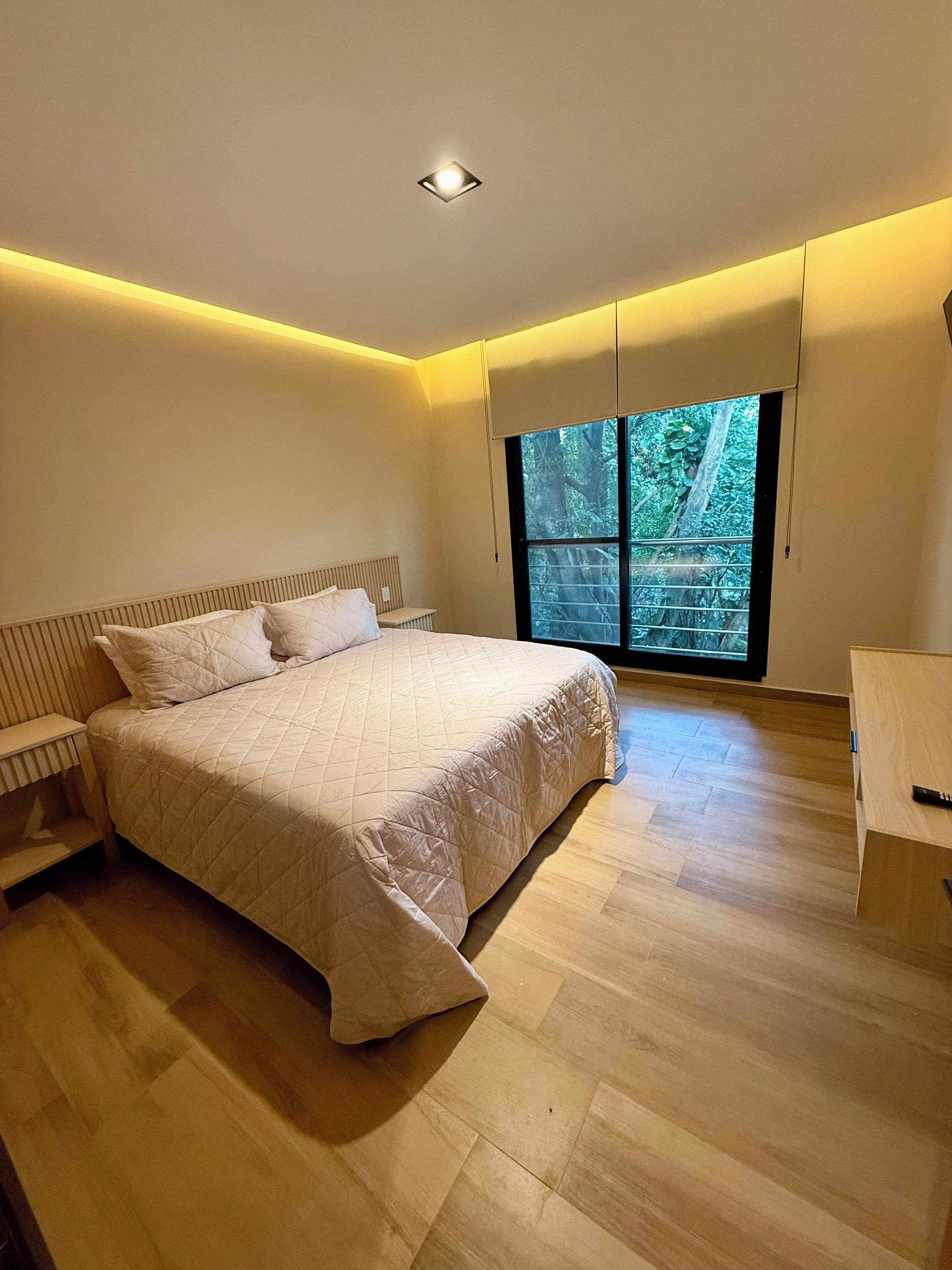 Un dormitorio moderno con una cama grande, una mesita de noche, un escritorio y una ventana con vista a zonas verdes.