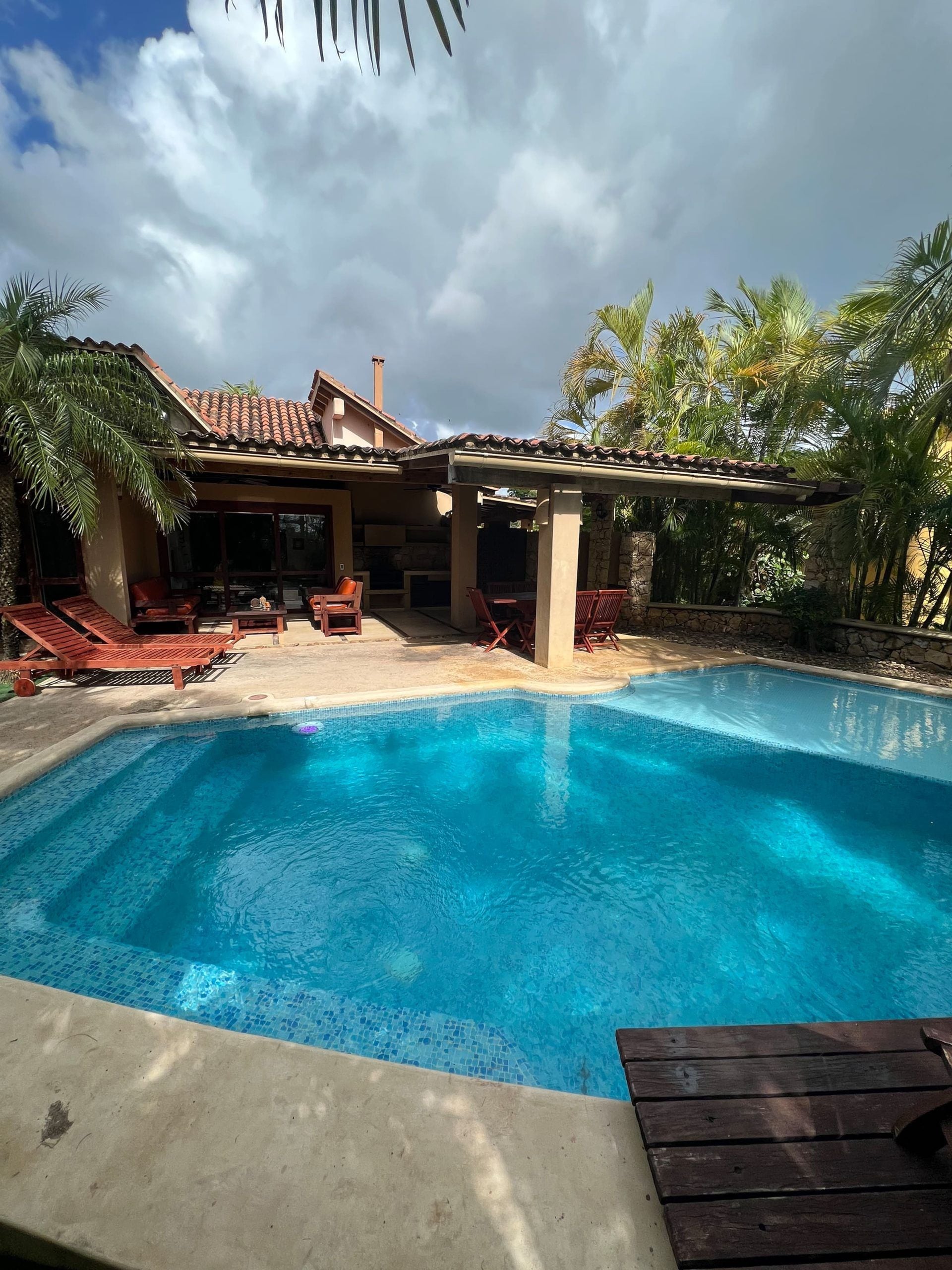 Una piscina con solárium de baldosas, sillas de descanso y un patio cubierto.