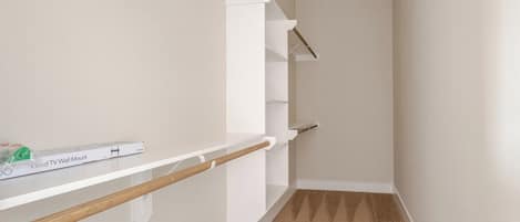 A narrow hallway with white walls and a carpeted floor. There are white shelves with wooden beams, and a box labeled 'TV Wall Mount' is visible on the left shelf.