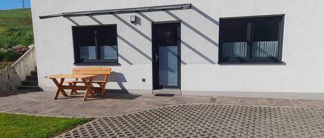 Ein modernes Gebäude mit weißer Fassade, zwei Fenstern, einer Tür und einer Holzbank vor der Eingangstür.