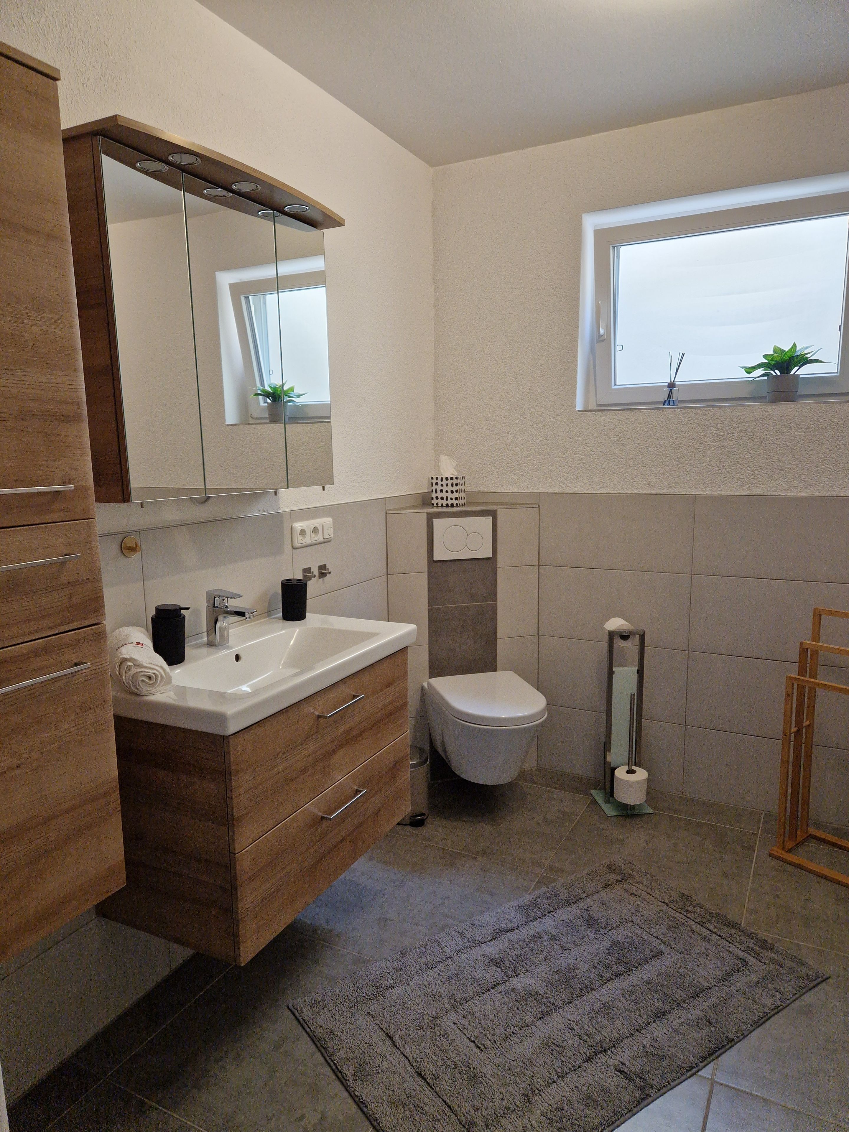 Ein modernes Badezimmer mit einem Holz-Waschtisch, einem weißen Waschbecken, einer Toilette und einem Spiegel über dem Waschtisch.