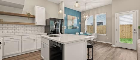 Una cocina moderna con muebles blancos, una isla central, iluminación colgante y vistas al patio trasero.