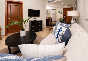 A living room with a sofa, a coffee table, a TV, a kitchen in the background, and a door. - Apartment with Ocean View in Puerto Bahía w/ AC and fast WIFI (Doncel)