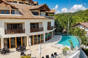 A multi-story hotel with a swimming pool, outdoor seating, and a view of a tropical landscape.
