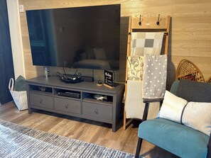 A modern living room with a flat-screen TV, a dark wooden entertainment unit, a small table with a decorative item, a sofa with cushions, and a patterned curtain.