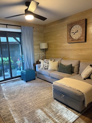 A living room with a ceiling fan, a clock, a sofa, a blue ottoman, and a view through a glass door.