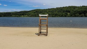 Un banc en bois sur une plage de sable, avec un lac et des collines boisées en arrière-plan.