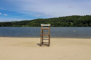 Een houten bankje op een zandstrand, met een meer en beboste heuvels op de achtergrond.