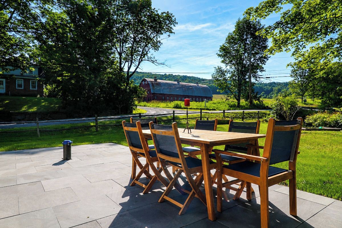 Een buitenpatio met een houten tafel en stoelen, een metalen prullenbak en uitzicht op een schuur en bomen.