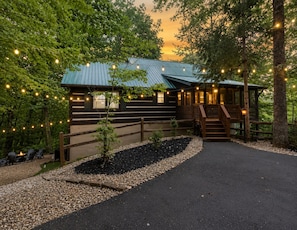 Un chalet rustique avec un toit en métal, entouré d’arbres et accessible par un chemin de gravier menant à l’entrée.