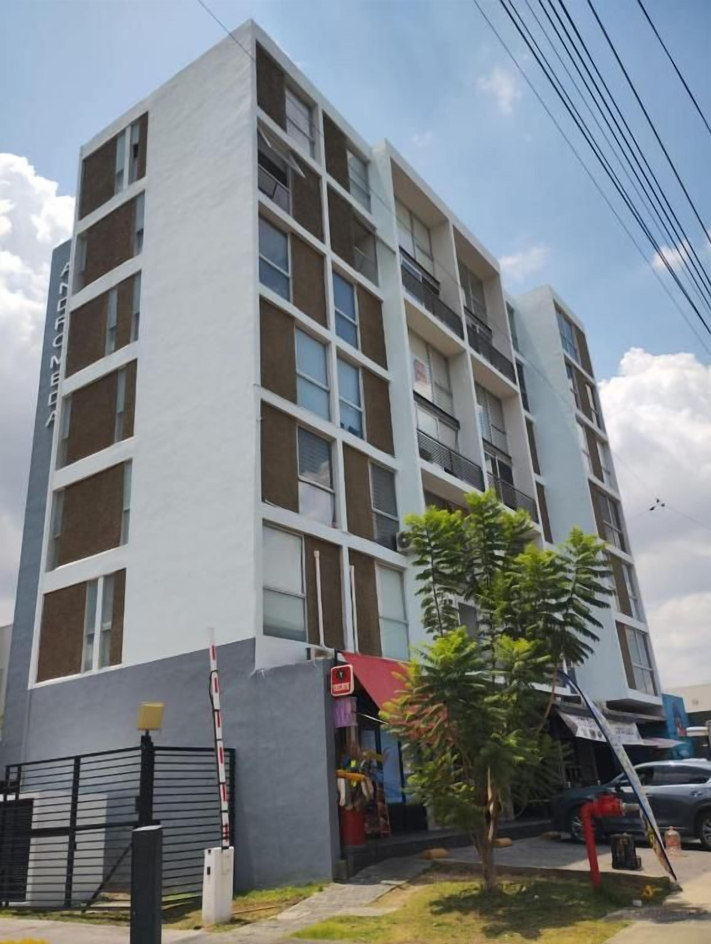 Un edificio de varios pisos con entrada protegida por un portón, un árbol pequeño y un balcón visible en los pisos superiores.