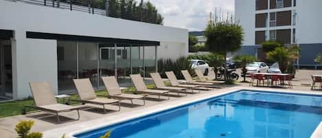 A swimming pool with lounge chairs, a covered area, and a building in the background.