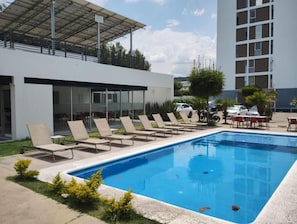 Una piscina con sillones de descanso, una zona cubierta y un edificio al fondo.
