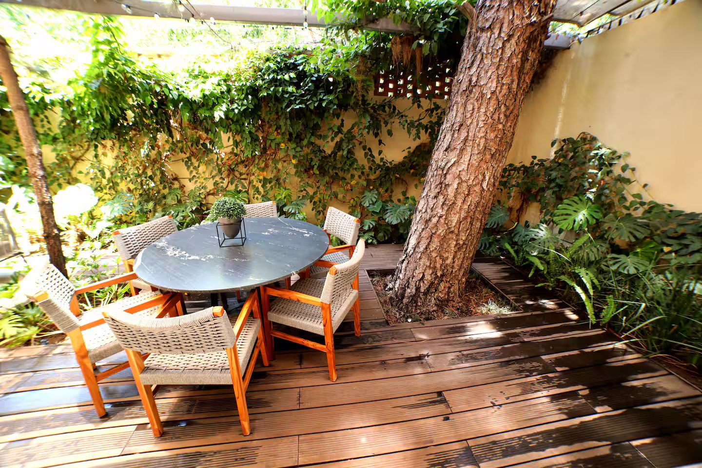 An outdoor patio with a round table and wicker chairs, surrounded by lush greenery and a tree.