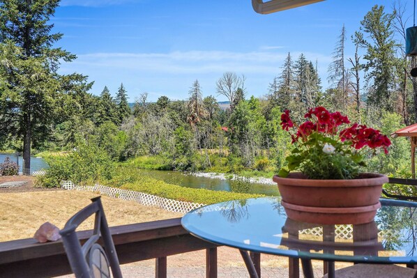 A patio with a round table and chairs, a potted plant with red flowers, and a view of a river and forested area.