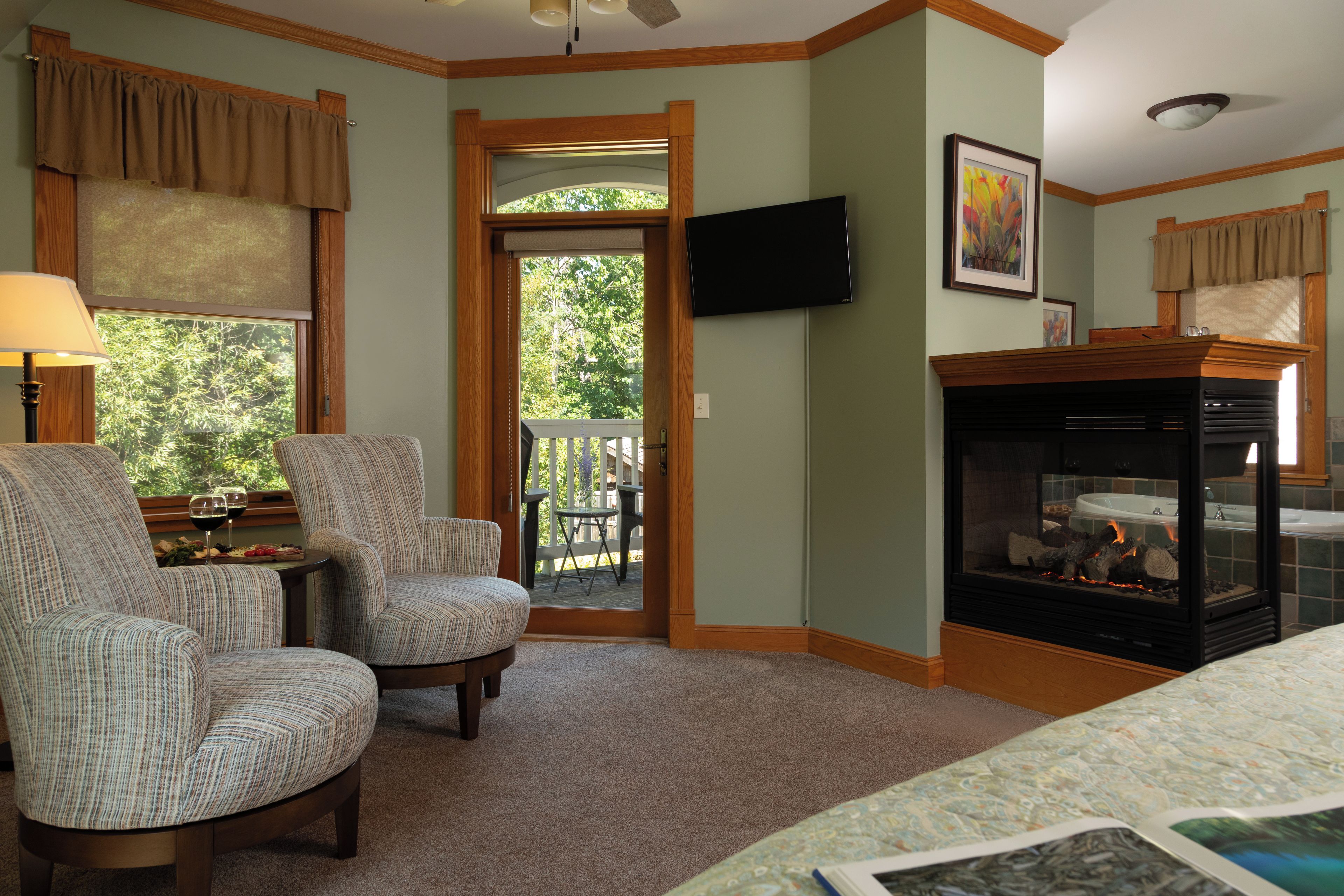 A room with two upholstered chairs, a small table with a glass of wine, a fireplace, and a television mounted on the wall.