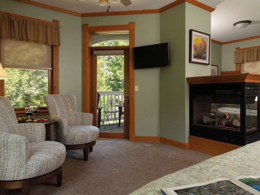 A room with two upholstered chairs, a small table with a glass of wine, a fireplace, and a television mounted on the wall.