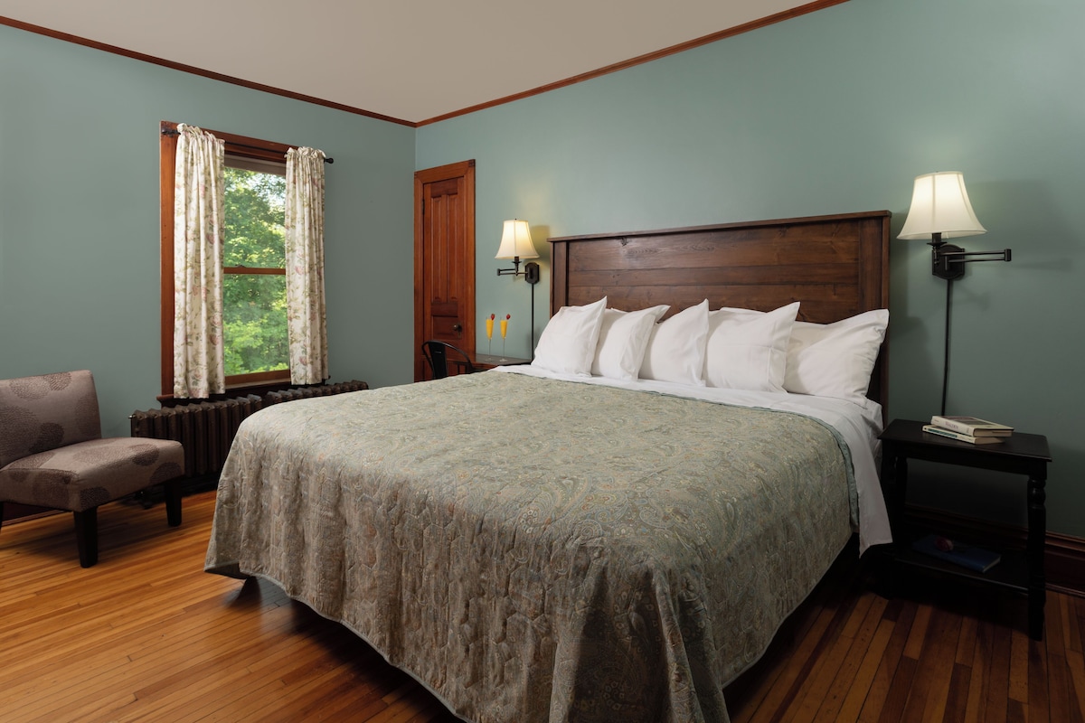 A bedroom with a large bed, two bedside lamps, a chair, and a window with curtains.