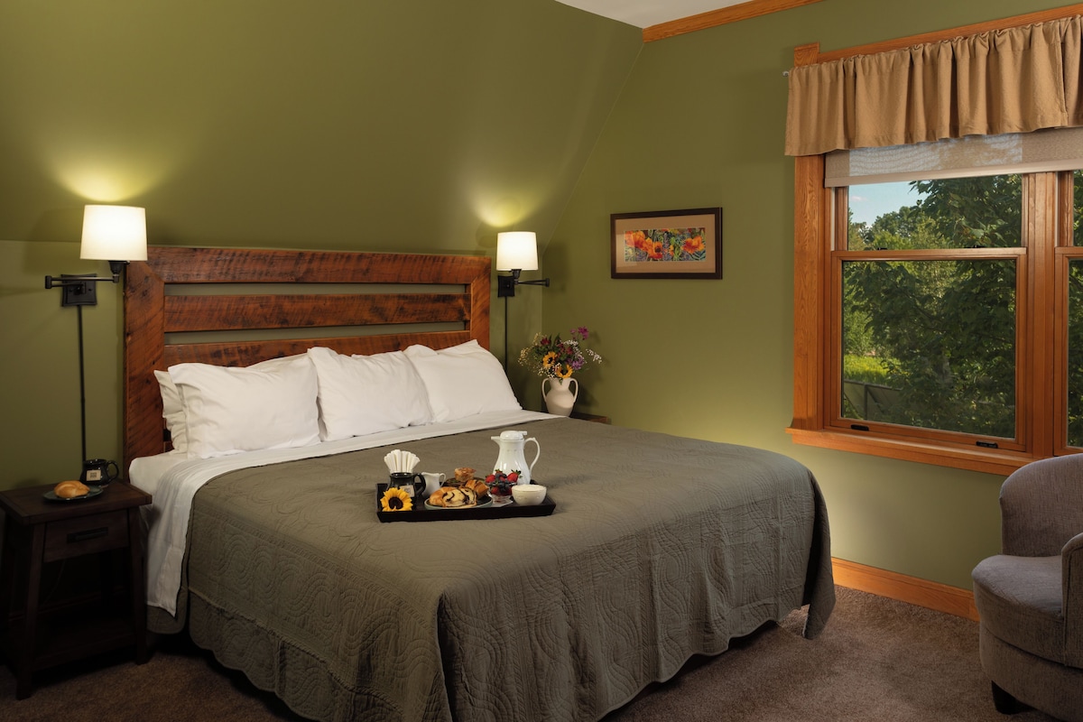 A neatly made bed with a tray of breakfast items, a nightstand with a lamp, and a window with a view of trees.