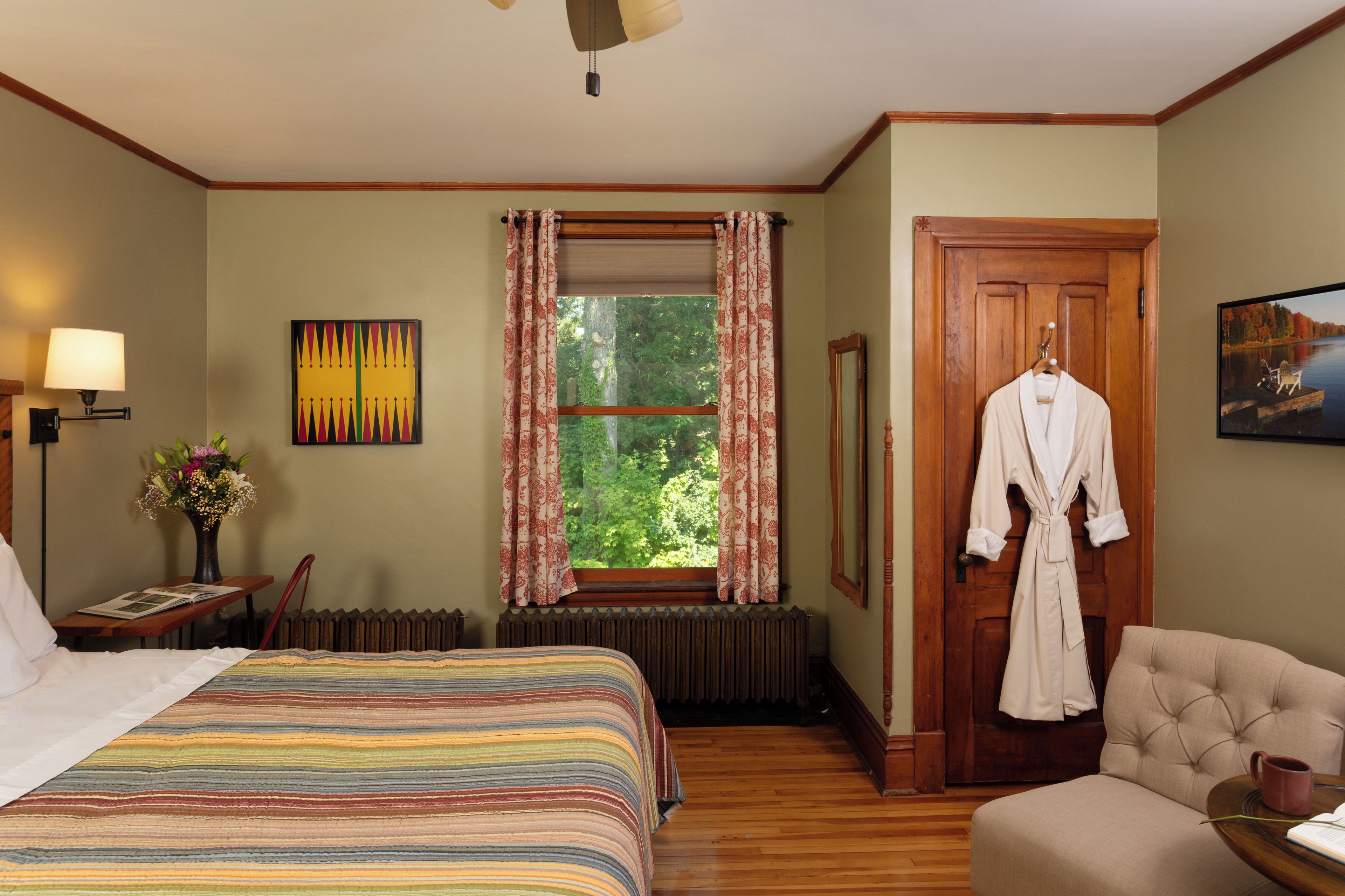 Een hotelkamer met een groot bed, een stoel, een houten vloer, een radiator, een raam met gordijnen, een lamp, een schilderij en een badjas die aan de deur hangt.