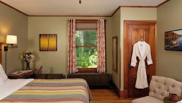 Een hotelkamer met een groot bed, een stoel, een houten vloer, een radiator, een raam met gordijnen, een lamp, een schilderij en een badjas die aan de deur hangt.