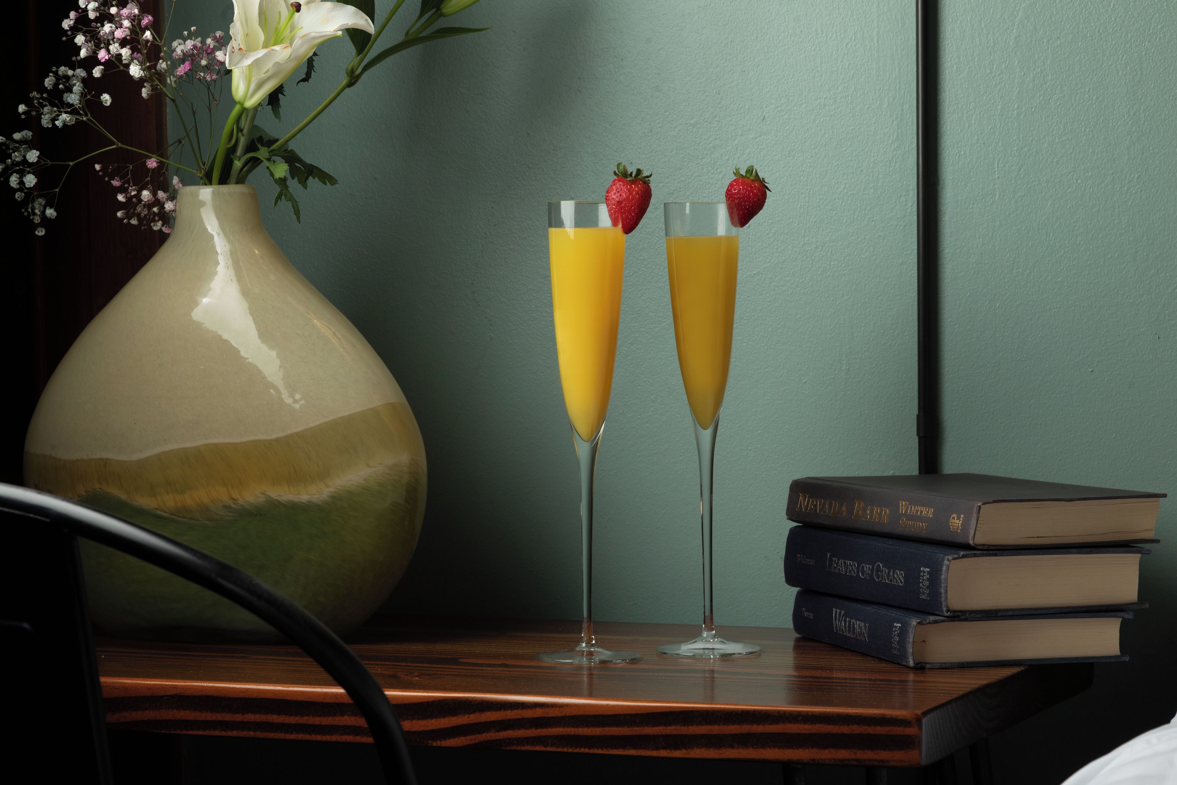 Deux verres de jus d’orange avec des fraises, posés sur une table, à côté d’un vase rempli de fleurs et d’une pile de livres.