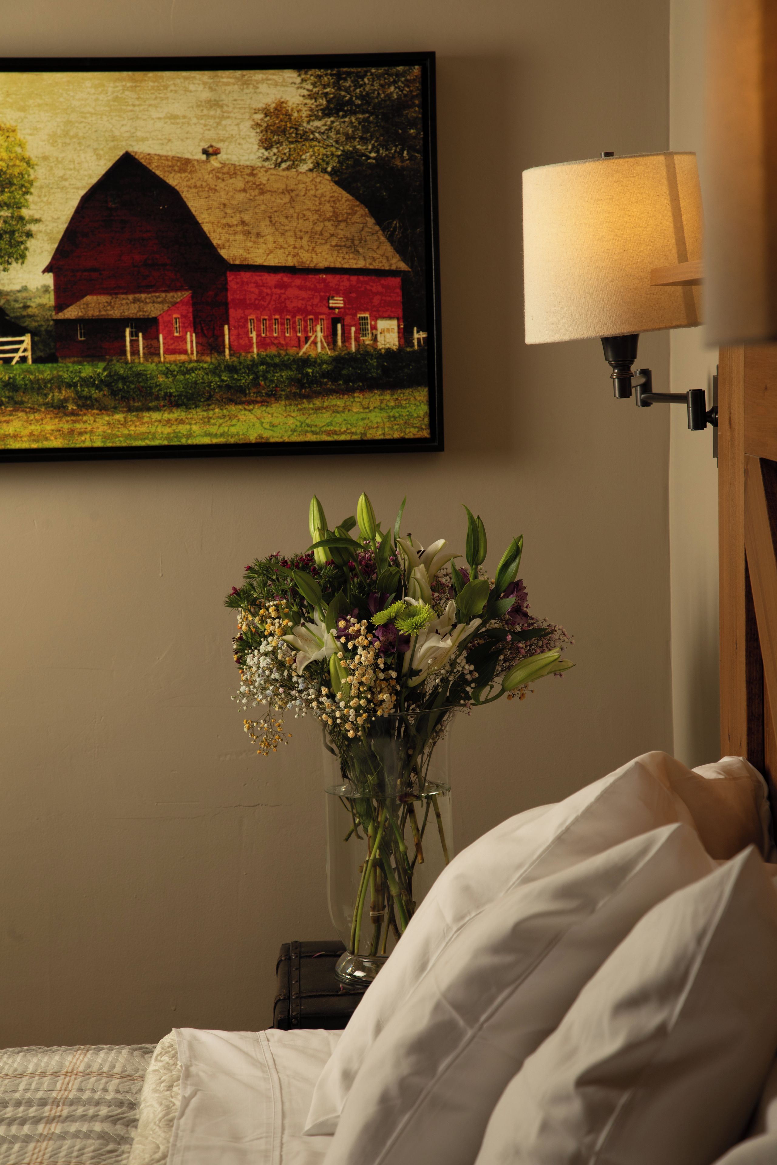 A bedroom with a bed, a wall-mounted lamp, a framed picture of a barn, and a vase with flowers.