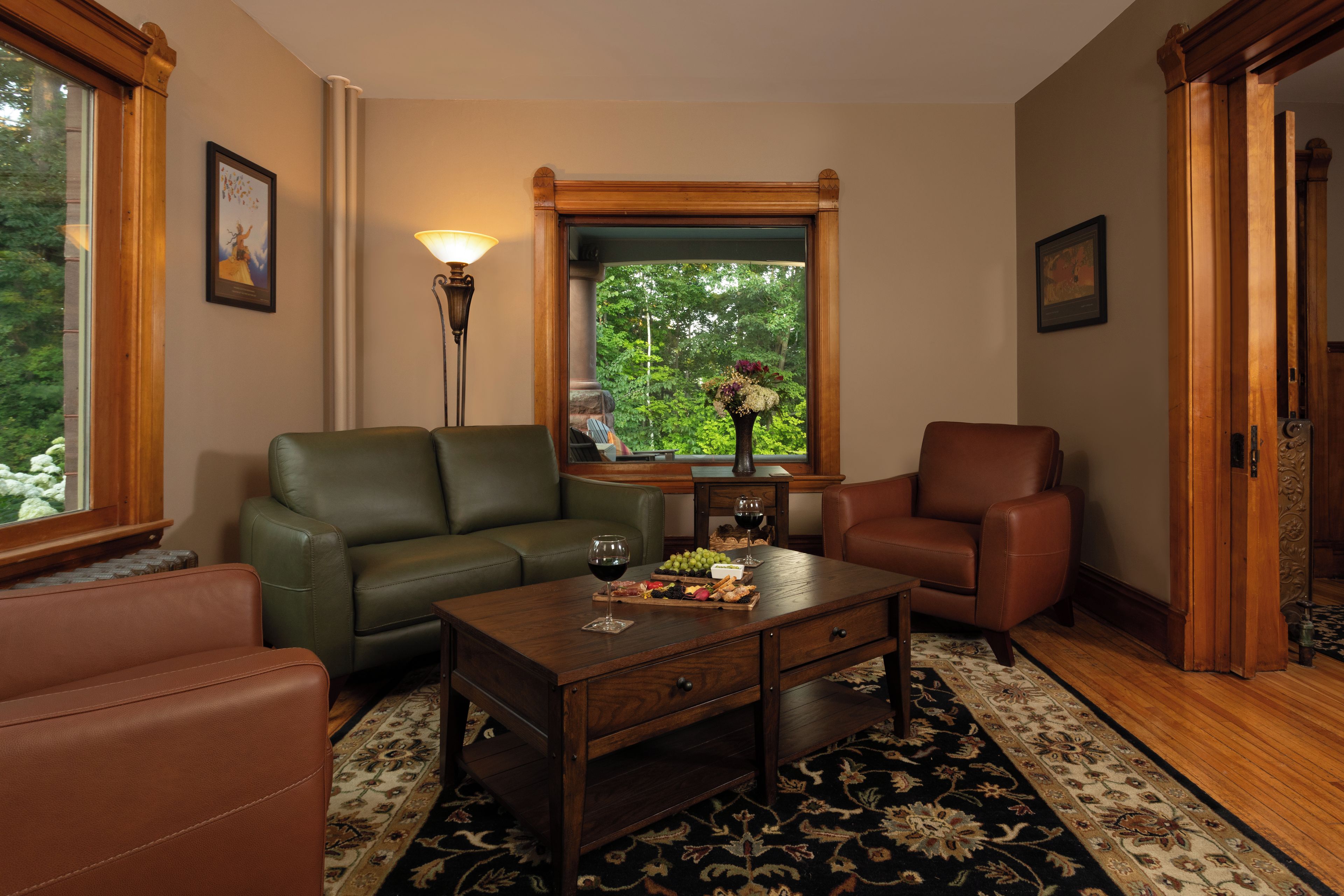 A living room with a green sofa, a brown leather armchair, a wooden coffee table, and a window with a view of trees.
