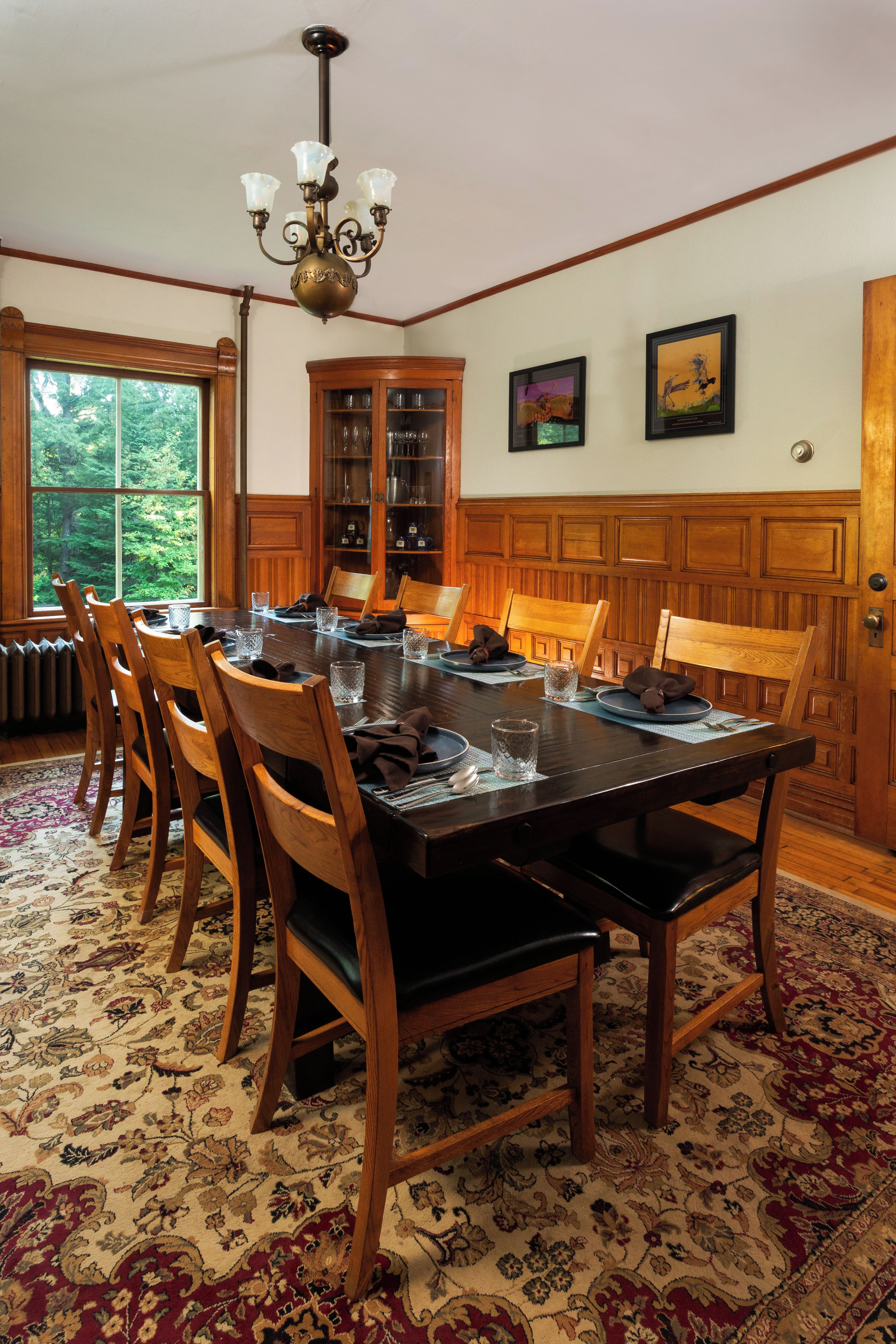 A dining room with a wooden table set for six, surrounded by chairs, with a chandelier above and a cabinet with glass doors to the side.
