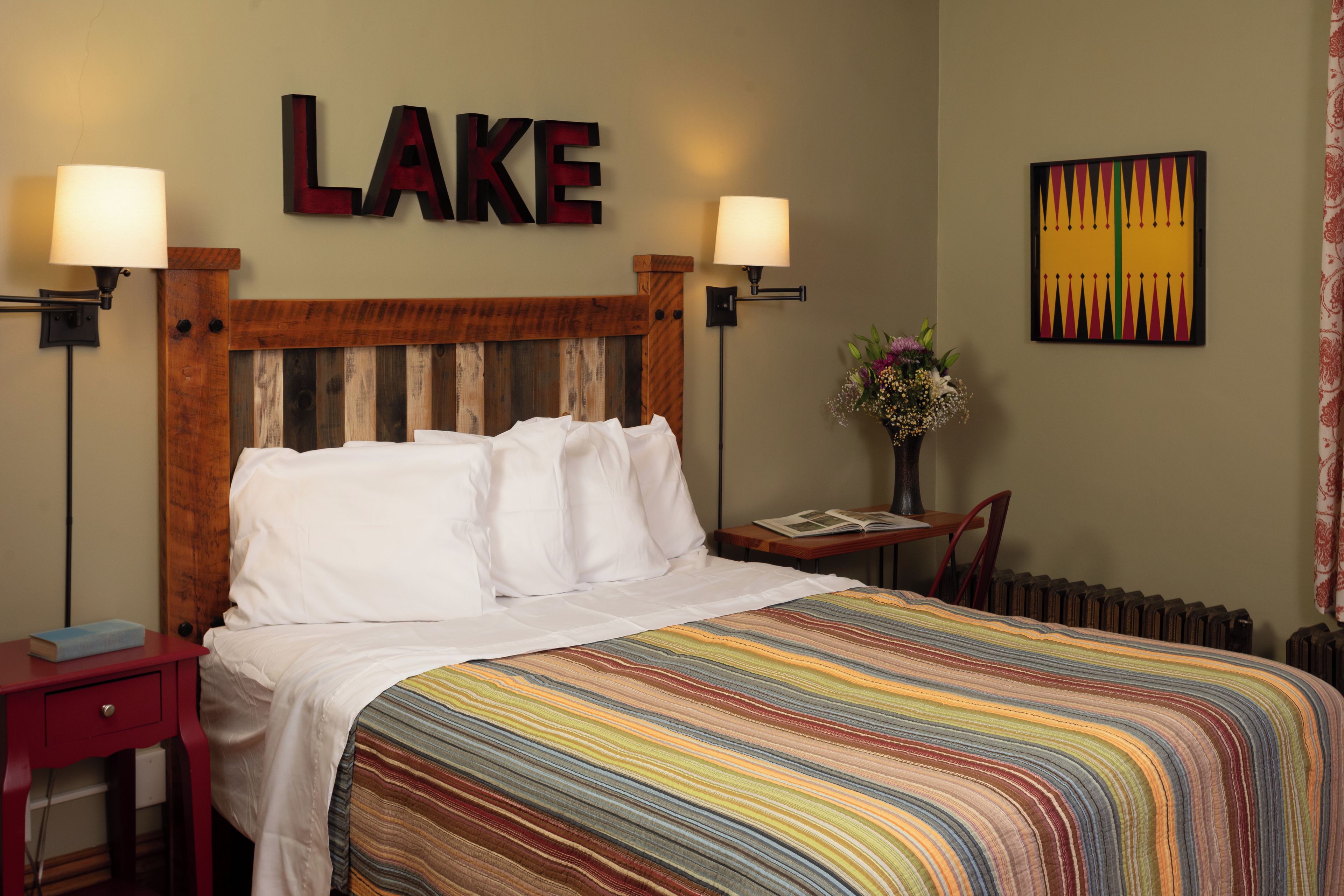 A hotel room with a bed, two bedside lamps, a nightstand, a chair, a table with a book, a vase with flowers, and a wall with a 'LAKE' sign and a colorful painting.