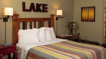 A hotel room with a bed, two bedside lamps, a nightstand, a chair, a table with a book, a vase with flowers, and a wall with a 'LAKE' sign and a colorful painting.