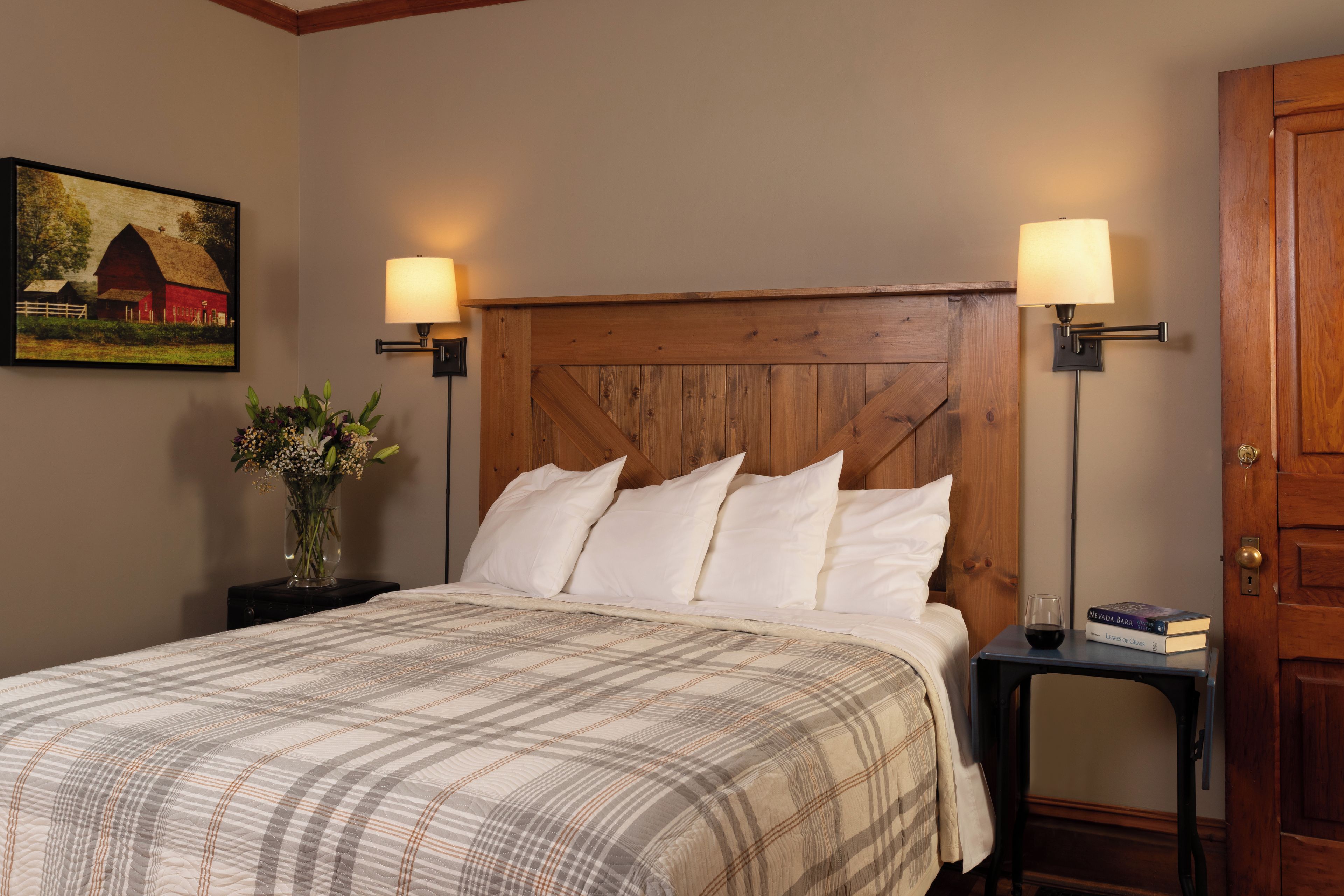 A neatly made bed with a plaid bedspread, flanked by two bedside tables with lamps, a vase of flowers, and a painting on the wall.
