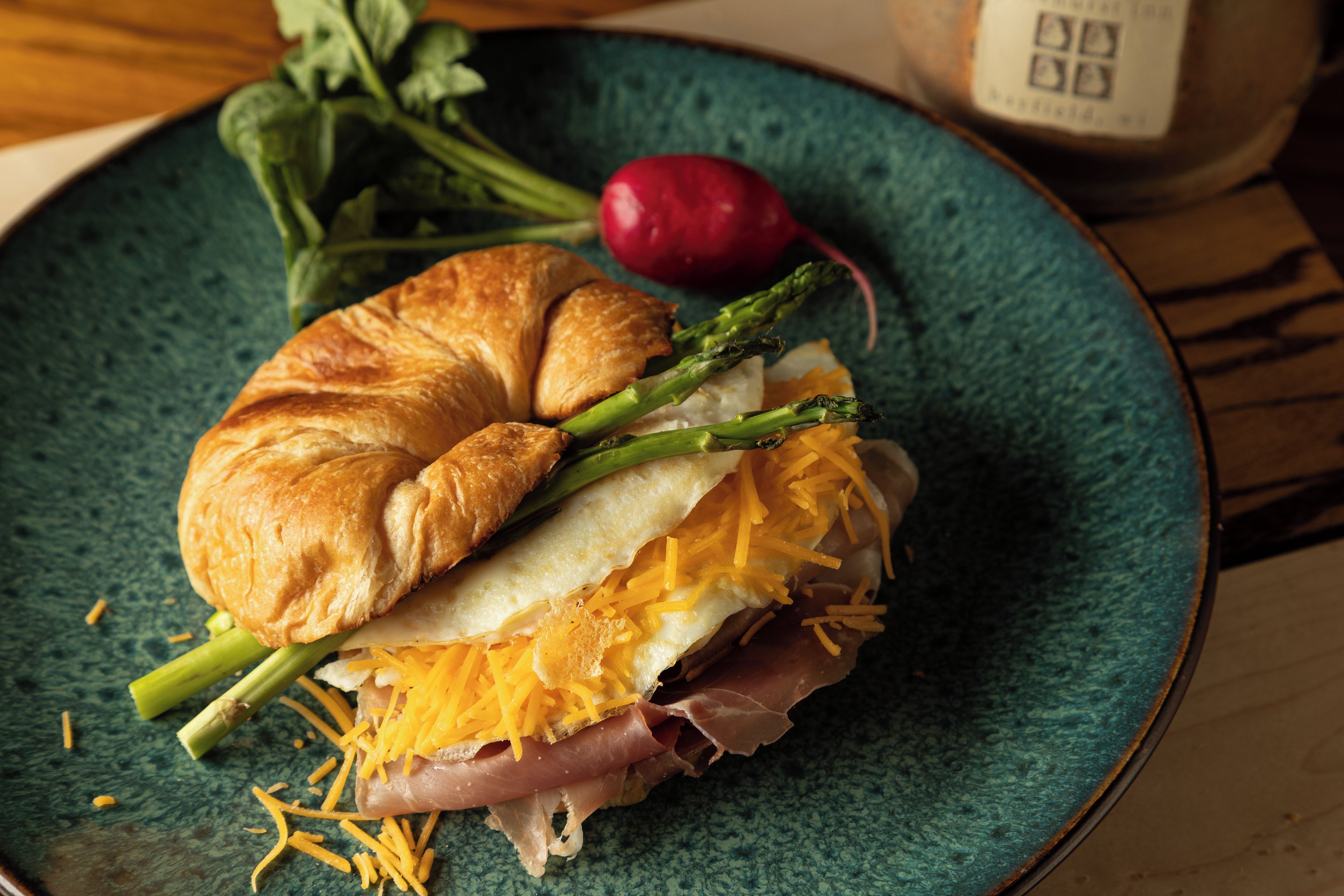 A sandwich with a croissant, egg, cheese, and meat, on a green plate with vegetables.