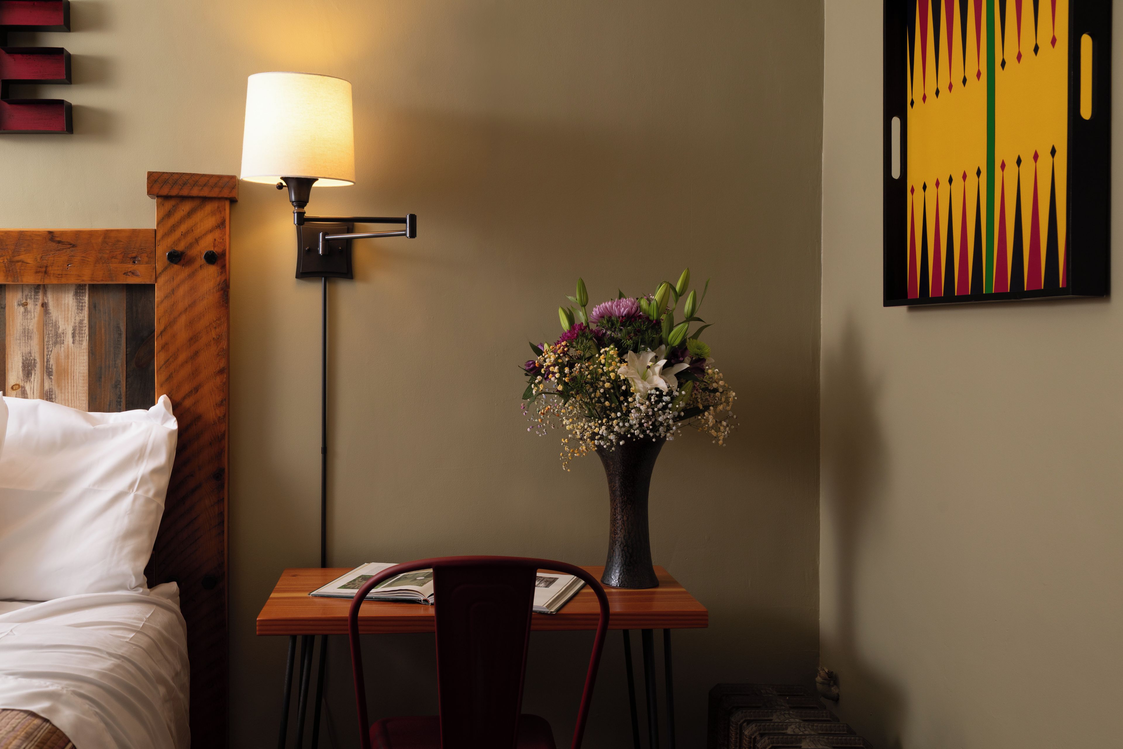 Ett hotellrum med en säng, ett skrivbord med en lampa, en vas med blommor och ett konstverk på väggen.