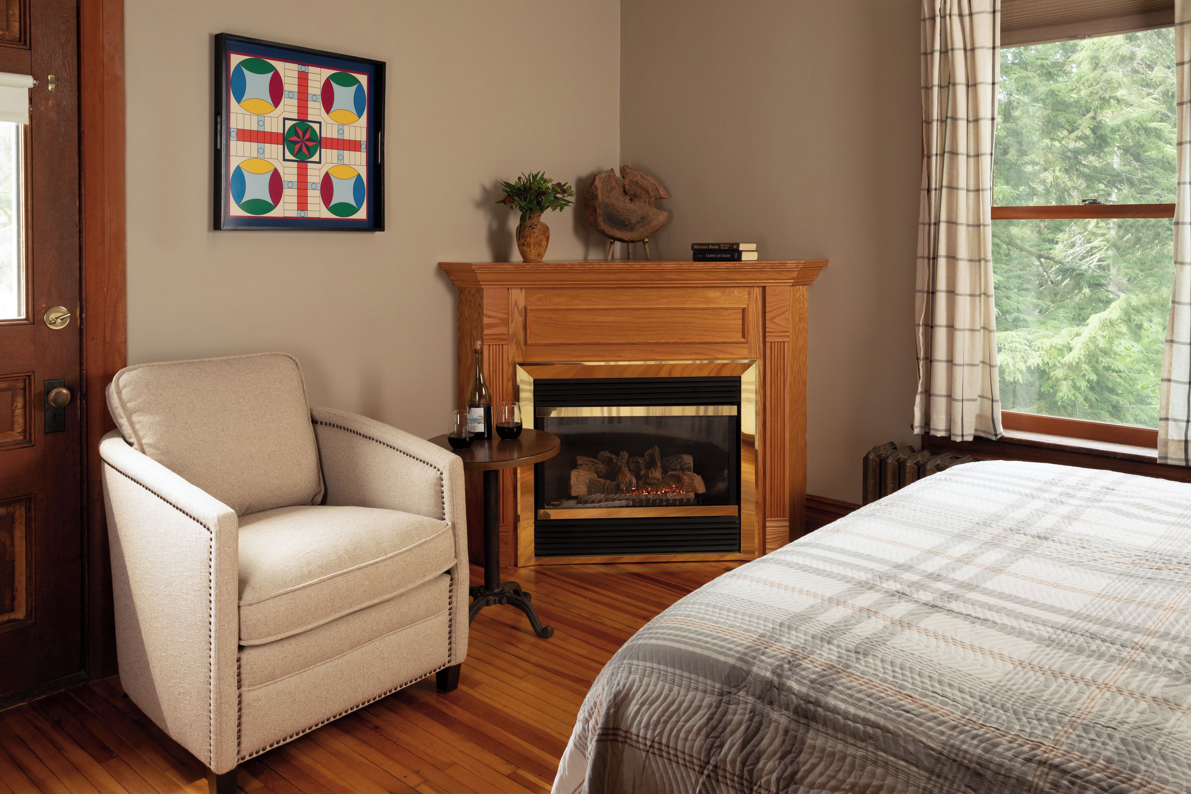 Quarto com piso de madeira, uma poltrona bege, lareira com manto e uma janela com cortinas.
