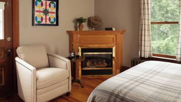 Quarto com piso de madeira, uma poltrona bege, lareira com manto e uma janela com cortinas.