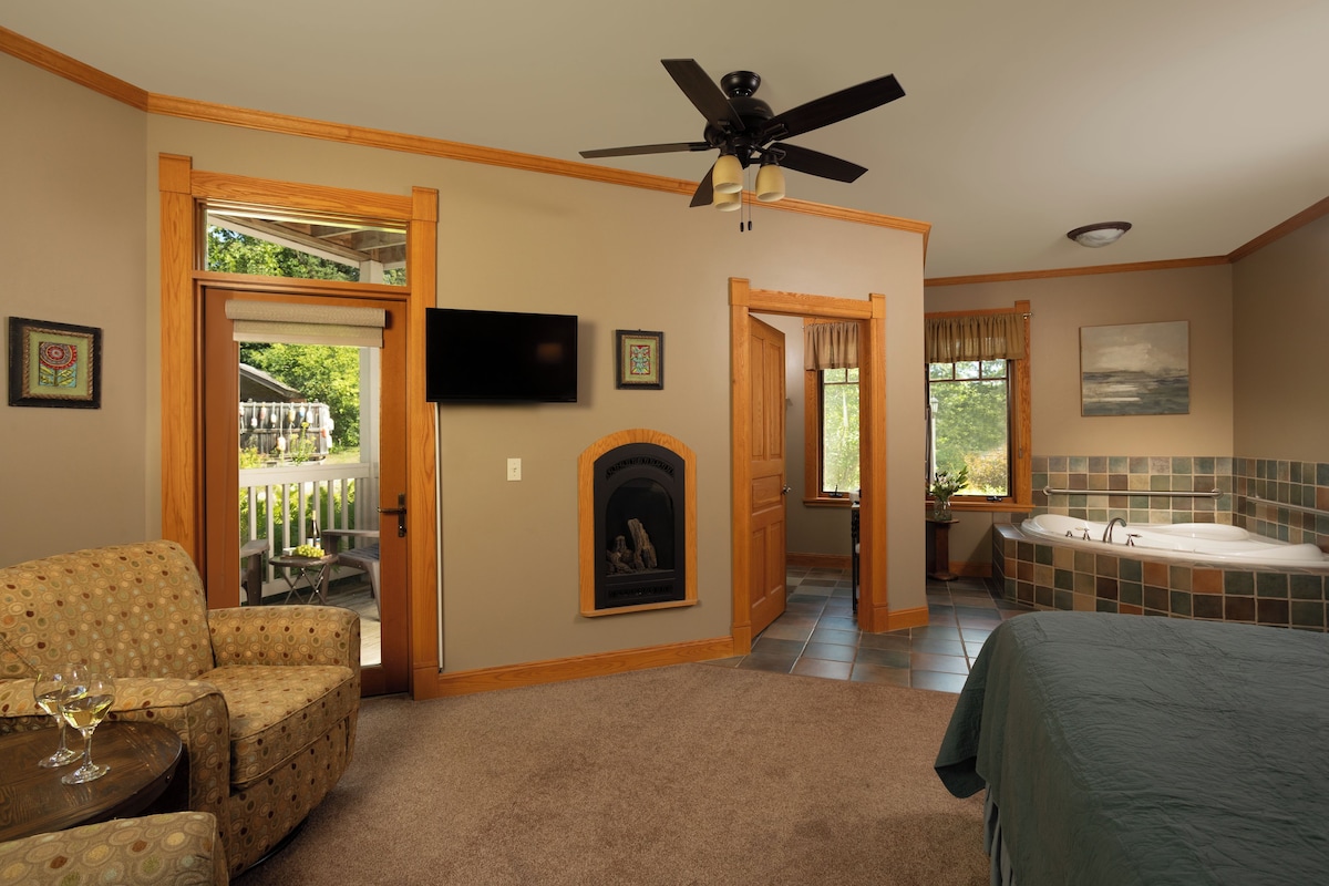 A bedroom with a bed, a ceiling fan, a fireplace, a TV, and a view of the outdoors.