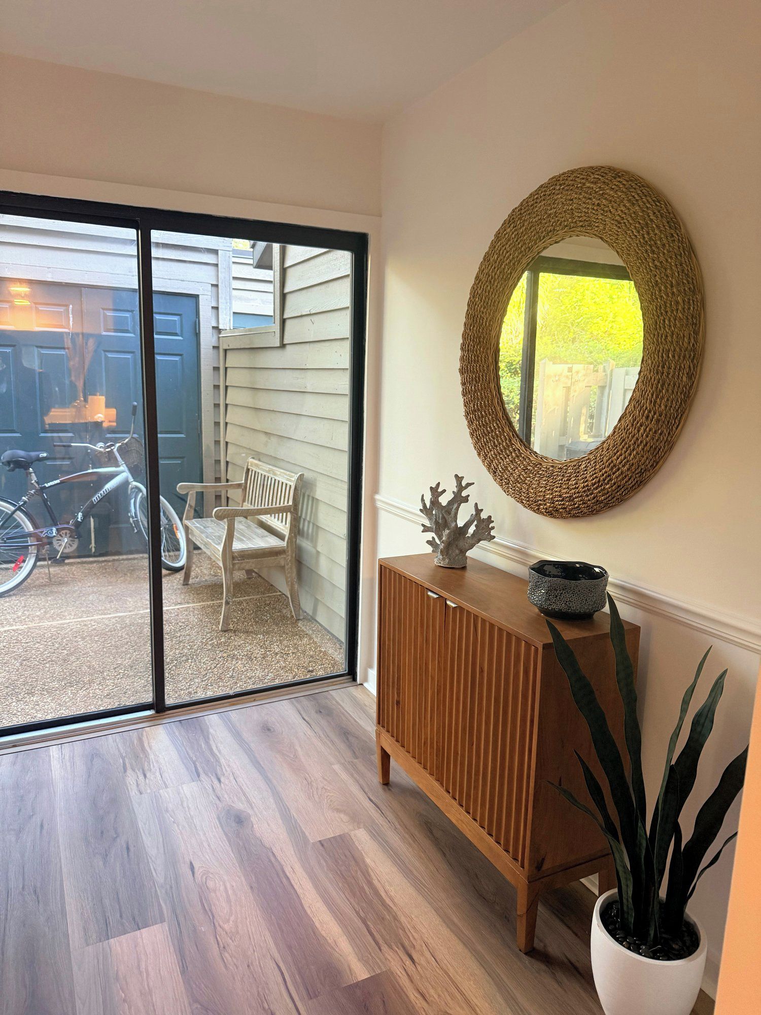 A room with a sliding glass door leading to a patio, a wooden cabinet, a round mirror, and a potted plant.