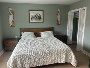 Ein Schlafzimmer mit einem großen Bett, zwei Nachttischen, einem Holzkopfteil, einem gerahmten Bild an der Wand und einer Tür zu einem anderen Raum.