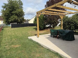 En træpergola med lyskæder, et overdækket udendørs bord og en græsplæne.
